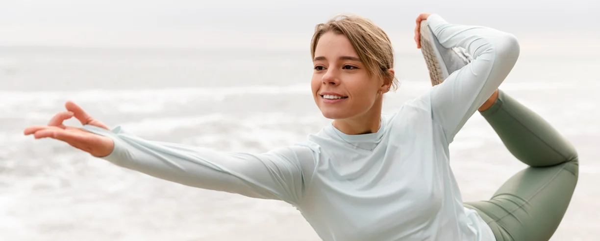 Een vrouw die yoga doet in een evenwichtige houding aan zee, waarmee ze flexibiliteit, mindfulness en buiten sporten promoot.